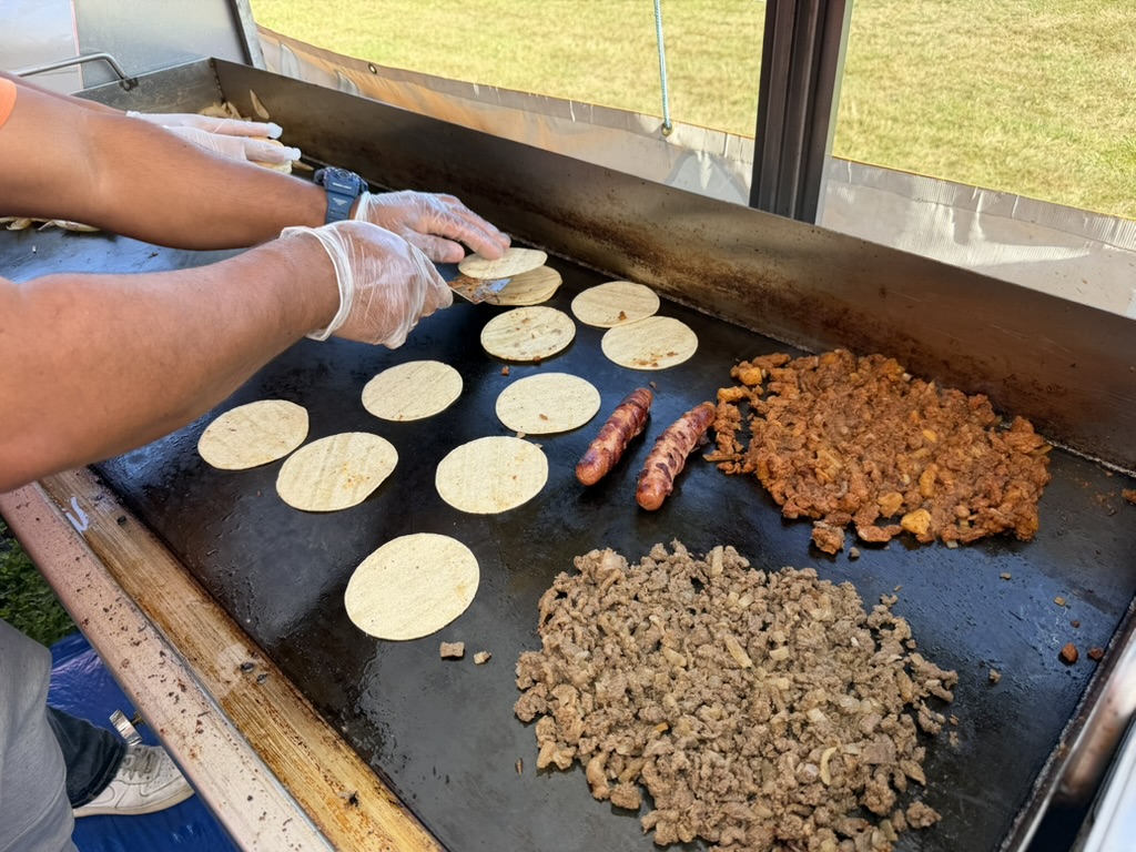 Tortillas, salchichas y dos tipos de carne en la plancha del puesto de Antojitos Corona durante un evento al aire libre