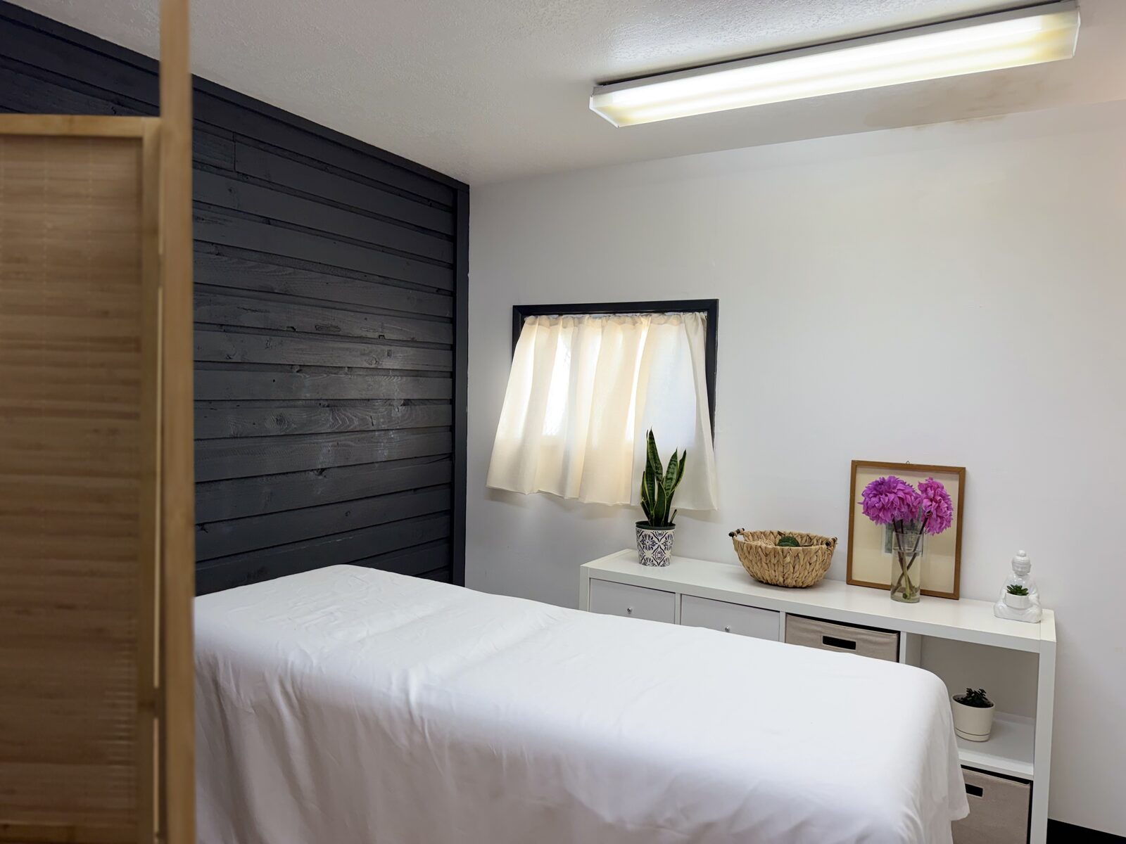 Sala de masaje de Casa Latido con camilla blanca, pared de madera oscura, cortina con luz suave y flores sobre un mueble