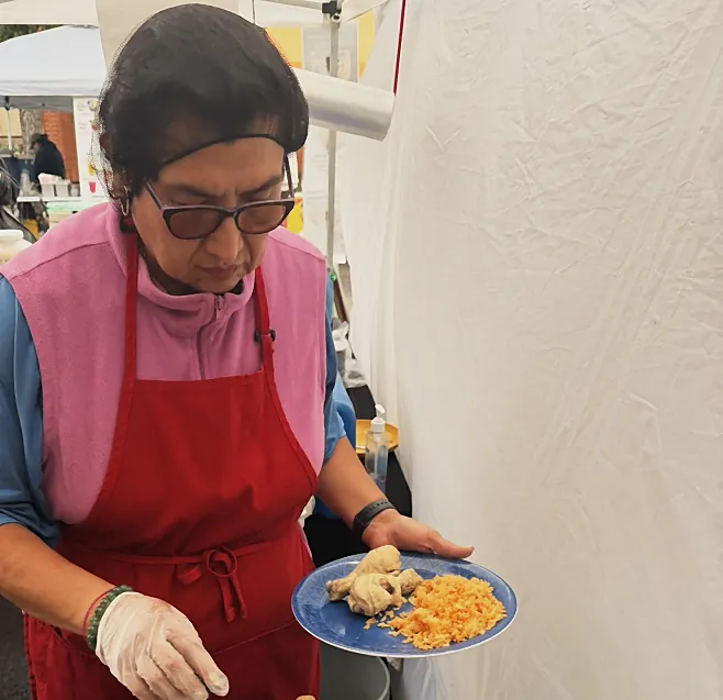 Una persona con delantal rojo sirve arroz y carne en un puesto de comida bajo una carpa.