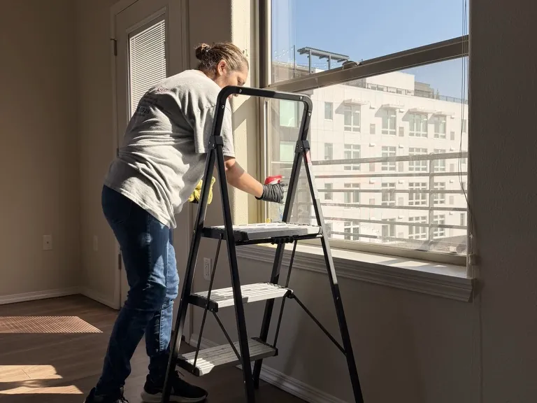 Una persona limpia una ventana grande desde una escalera en un departamento con luz de la tarde.